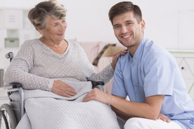 Positive senior woman on a wheelchair and smiling male carer, light home interior