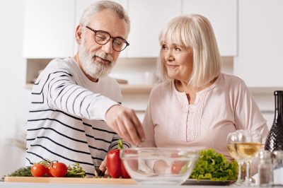 Enjoying healthy nutrition. Lively careful upbeat senior couple standing in the kitchen and cooking healthy dinner while expressing care and love
