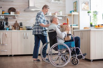 Cheerful wife helping disabled husband in the kitchen. Senior woman taking grocery paper bag from handicapped husband in wheelchair. Mature people with fresh vegetables from market. Living with disabled person with walking disabilities