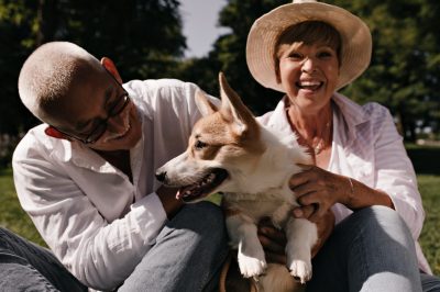 Grey haired man in eyeglasses and white shirt smiling and posing with cute corgi and blonde woman in hat and light blouse outdoor..